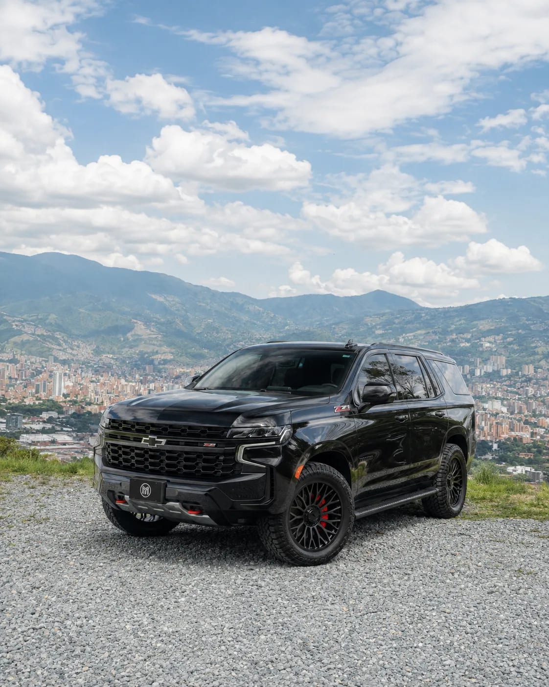 Black Chevrolet Tahoe with dark wheels, front three-quarter view, at a lookout with Medellín and the mountains behind.