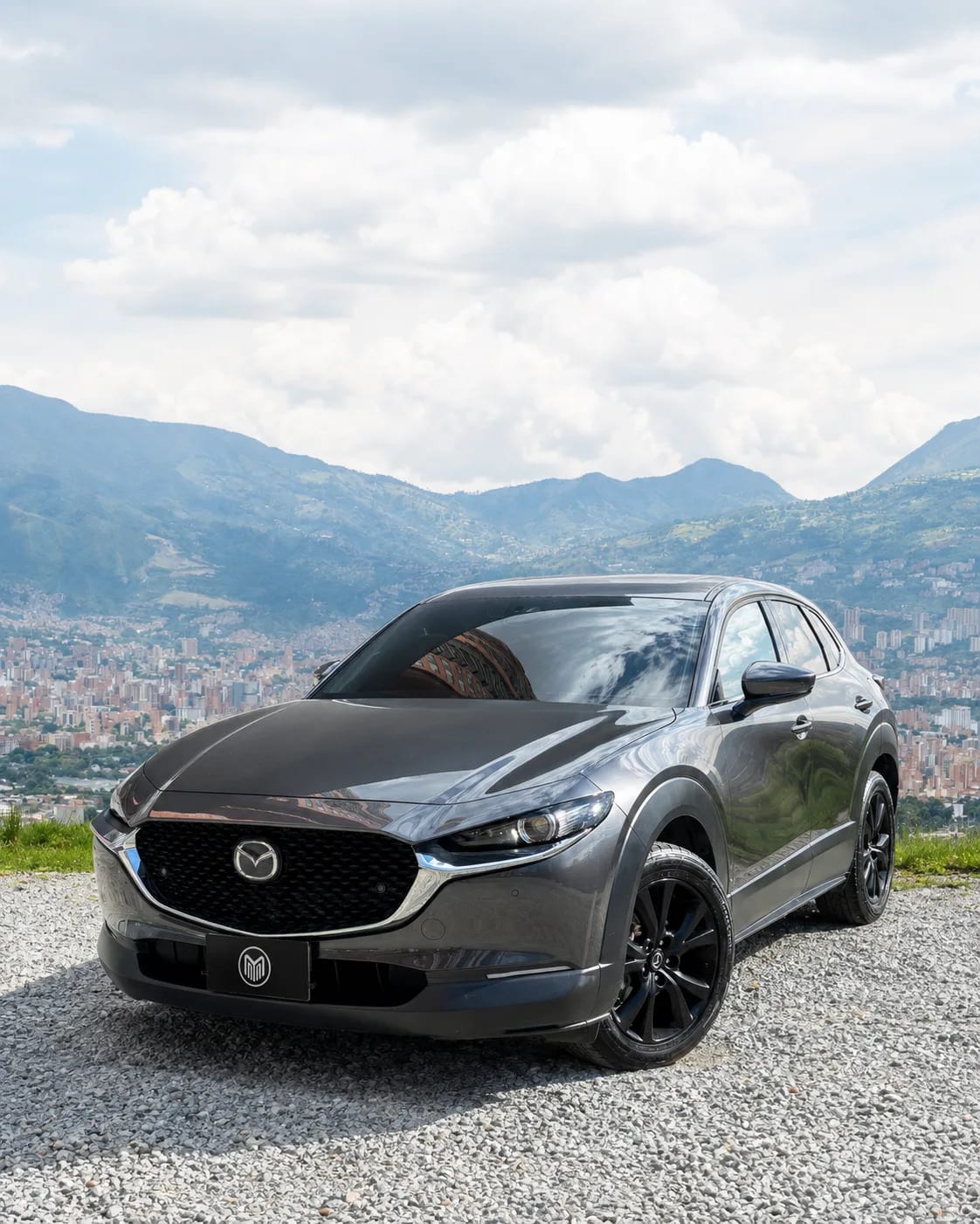 Mazda CX-30 gris grafito con rines negros, de tres cuartos frontal, sobre un mirador con Medellín al fondo.